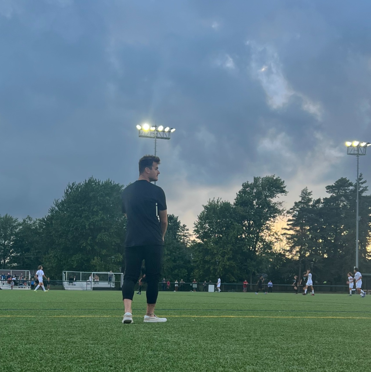 Coach Yohan Andraud standing on the field at dusk under floodlights.
